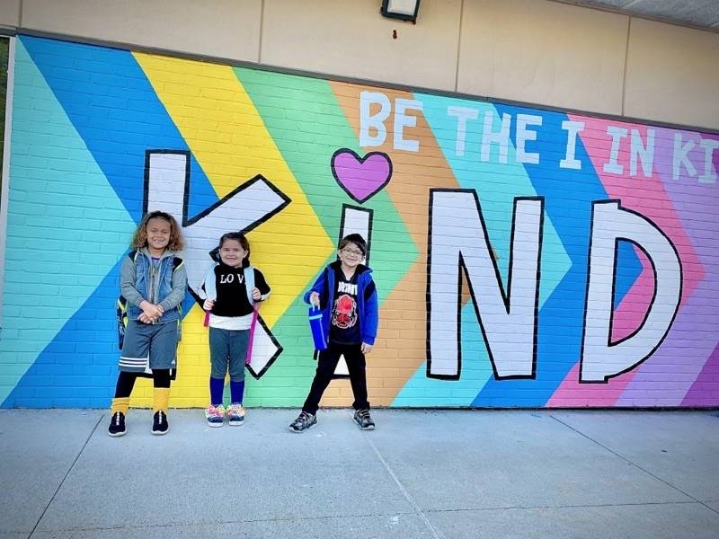 Students posing in front of a wall mural saying "Be the I in Kind!"