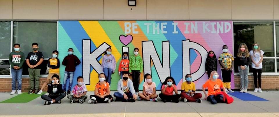 Students posing in front of a wall mural that says "kind"